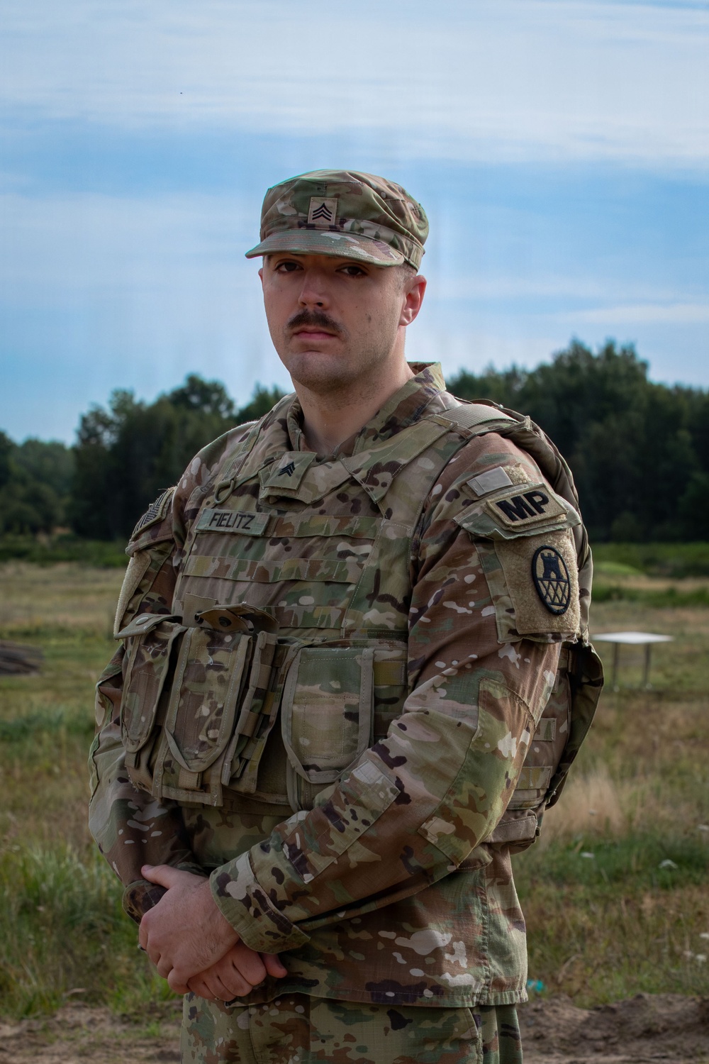 A U.S. Army Military Police sergeant stands at parade rest in an outdoor field environment. The soldier is wearing OCP (Operational Camouflage Pattern) uniform with a plate carrier vest and multiple visible unit and branch insignia. The setting appears to be a training range or field exercise area.