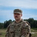 A U.S. Army Military Police sergeant stands at parade rest in an outdoor field environment. The soldier is wearing OCP (Operational Camouflage Pattern) uniform with a plate carrier vest and multiple visible unit and branch insignia. The setting appears to be a training range or field exercise area.