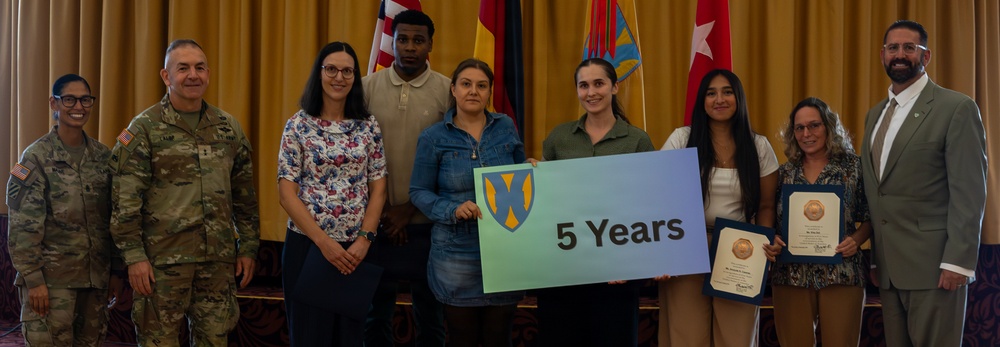 A group of approximately nine people pose for a formal recognition photo in what appears to be a military or government ceremony setting. Two individuals in U.S. Army combat uniforms stand alongside civilians, with one person holding a sign reading '5 Years' featuring a shield emblem. Several participants are holding certificates or awards, suggesting a service anniversary recognition event.