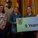 A group of approximately nine people pose for a formal recognition photo in what appears to be a military or government ceremony setting. Two individuals in U.S. Army combat uniforms stand alongside civilians, with one person holding a sign reading '5 Years' featuring a shield emblem. Several participants are holding certificates or awards, suggesting a service anniversary recognition event.