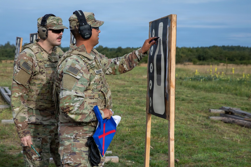Two U.S. Army soldiers in OCP (Operational Camouflage Pattern) uniforms are examining a shooting target on a range. One soldier, identified by name tape as 'SOLTANO', is pointing at the target while holding a blue flag with a red cross, while the other soldier identified as 'FIELITZ' observes. Both are wearing hearing protection and appear to be conducting a post-shooting assessment.