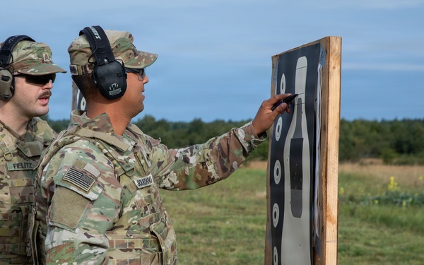 Two U.S. Army soldiers in OCP (Operational Camouflage Pattern) uniforms are examining a shooting target on a range. One soldier, identified by name tape as 'SOLTANO', is pointing at the target while holding a blue flag with a red cross, while the other soldier identified as 'FIELITZ' observes. Both are wearing hearing protection and appear to be conducting a post-shooting assessment.