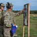 Two U.S. Army soldiers in OCP (Operational Camouflage Pattern) uniforms are examining a shooting target on a range. One soldier, identified by name tape as 'SOLTANO', is pointing at the target while holding a blue flag with a red cross, while the other soldier identified as 'FIELITZ' observes. Both are wearing hearing protection and appear to be conducting a post-shooting assessment.