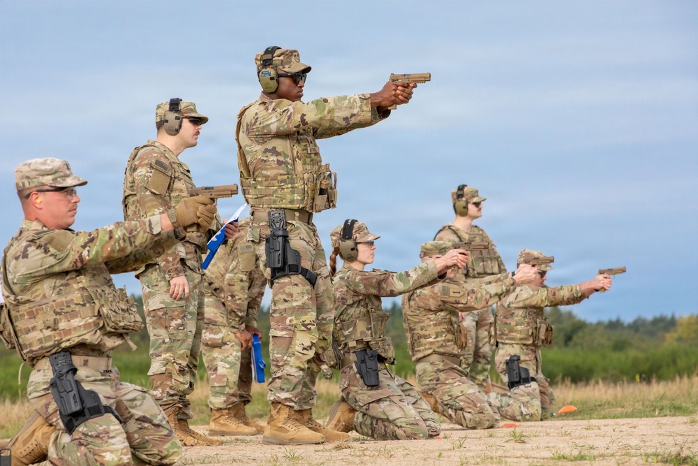A group of military personnel in MultiCam/OCP camouflage uniforms are conducting pistol qualification or training on an outdoor firing range. Soldiers are positioned in both standing and kneeling stances, aiming semi-automatic pistols downrange. The scene appears to be a supervised range exercise with an instructor or range officer demonstrating proper technique.