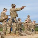A group of military personnel in MultiCam/OCP camouflage uniforms are conducting pistol qualification or training on an outdoor firing range. Soldiers are positioned in both standing and kneeling stances, aiming semi-automatic pistols downrange. The scene appears to be a supervised range exercise with an instructor or range officer demonstrating proper technique.