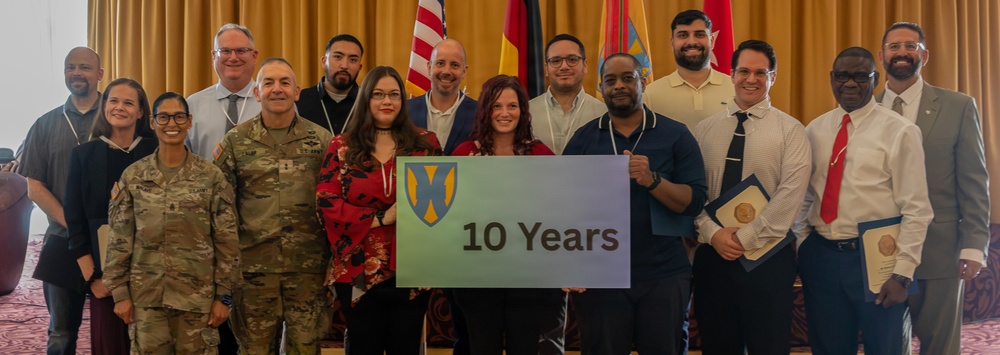 A group of approximately 15 people pose together for a formal group photo, holding a sign that reads '10 Years' featuring a military unit shield/crest logo. The setting appears to be an indoor venue with curtains and flags in the background, including the American flag and what appears to be a German flag. The occasion appears to be a 10th anniversary celebration, likely of a military program or unit.