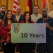 A group of approximately 15 people pose together for a formal group photo, holding a sign that reads '10 Years' featuring a military unit shield/crest logo. The setting appears to be an indoor venue with curtains and flags in the background, including the American flag and what appears to be a German flag. The occasion appears to be a 10th anniversary celebration, likely of a military program or unit.
