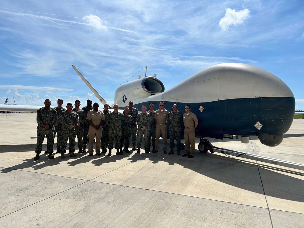 A group of approximately 15-16 U.S. Navy personnel in uniform are posing for a group photograph on a tarmac in front of a large unmanned aerial vehicle (UAV), identifiable as a Northrop Grumman MQ-4C Triton based on its distinctive fuselage shape, blue and white color scheme, and naval insignia. The aircraft is parked on a concrete flight line under partly cloudy skies. Personnel are wearing a mix of Navy Working Uniform (NWU) camouflage and khaki service uniforms.