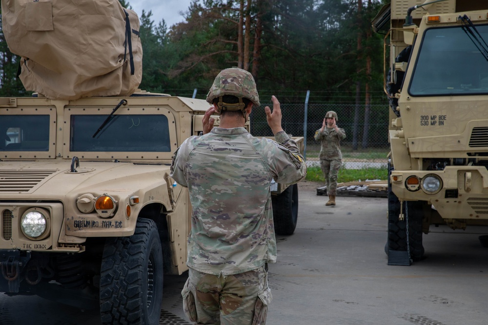 Two soldiers in U.S. Army OCP (Operational Camouflage Pattern) uniforms are conducting vehicle marshaling operations between military vehicles. The foreground soldier is using hand signals to communicate with a second soldier in the background, who is mirroring or responding with their own hand signals. The scene takes place in what appears to be a military motor pool or staging area with tan-colored tactical vehicles.
