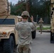 Two soldiers in U.S. Army OCP (Operational Camouflage Pattern) uniforms are conducting vehicle marshaling operations between military vehicles. The foreground soldier is using hand signals to communicate with a second soldier in the background, who is mirroring or responding with their own hand signals. The scene takes place in what appears to be a military motor pool or staging area with tan-colored tactical vehicles.