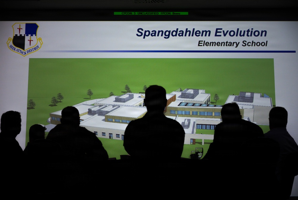 A group of silhouetted individuals are viewing a projected presentation slide titled 'Spangdahlem Evolution - Elementary School,' which displays an architectural rendering of a planned elementary school building. The presentation appears to be associated with Spangdahlem Air Base, as indicated by the military unit shield bearing the motto 'Seek Attack Destroy' in the upper left corner. The slide is classified as 'UNCLASSIFIED' as noted in the green banner at the top of the projection.