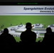 A group of silhouetted individuals are viewing a projected presentation slide titled 'Spangdahlem Evolution - Elementary School,' which displays an architectural rendering of a planned elementary school building. The presentation appears to be associated with Spangdahlem Air Base, as indicated by the military unit shield bearing the motto 'Seek Attack Destroy' in the upper left corner. The slide is classified as 'UNCLASSIFIED' as noted in the green banner at the top of the projection.