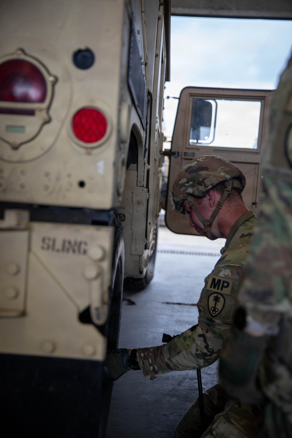 A U.S. Army Military Police (MP) soldier in OCP (Operational Camouflage Pattern) uniform and combat helmet is performing maintenance or an inspection on a tan/desert-colored HMMWV (Humvee) military vehicle. The soldier is crouched near the lower side of the vehicle, working with a tool or component near the wheel area. The setting appears to be an indoor motor pool or vehicle bay.