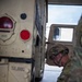 A U.S. Army Military Police (MP) soldier in OCP (Operational Camouflage Pattern) uniform and combat helmet is performing maintenance or an inspection on a tan/desert-colored HMMWV (Humvee) military vehicle. The soldier is crouched near the lower side of the vehicle, working with a tool or component near the wheel area. The setting appears to be an indoor motor pool or vehicle bay.