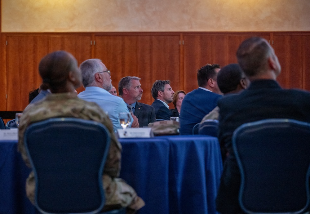 A formal meeting or conference is taking place in what appears to be a banquet or conference room. Multiple attendees, including at least one person in military camouflage uniform and several in business attire, are seated at round tables covered with blue tablecloths. The participants appear to be attentively watching a presentation or speaker located off-camera.