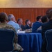 A formal meeting or conference is taking place in what appears to be a banquet or conference room. Multiple attendees, including at least one person in military camouflage uniform and several in business attire, are seated at round tables covered with blue tablecloths. The participants appear to be attentively watching a presentation or speaker located off-camera.