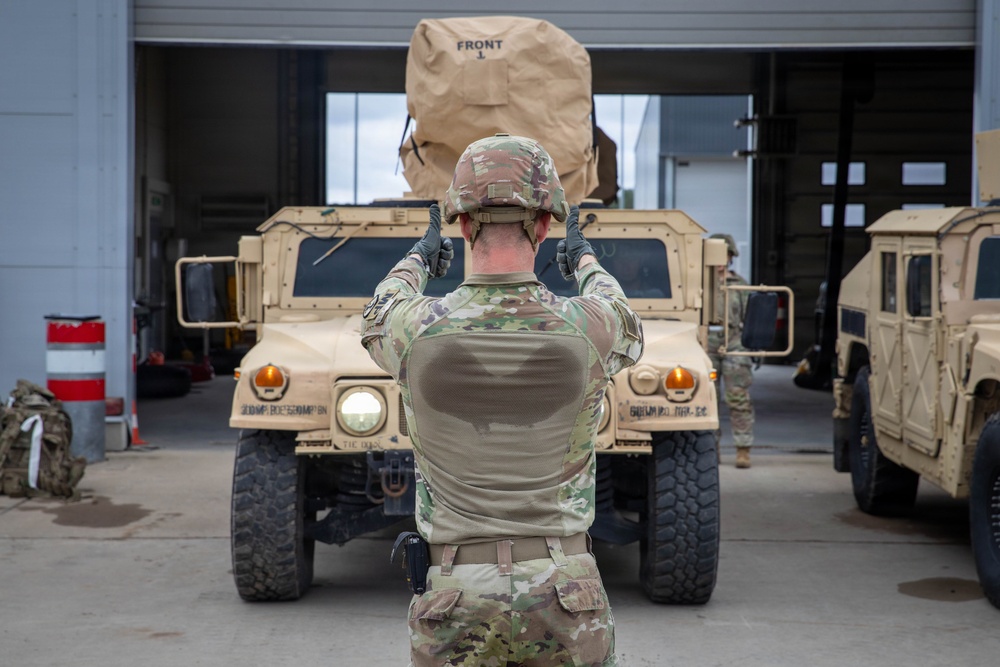 A U.S. Army soldier in OCP (Operational Camouflage Pattern) uniform is giving hand signals with both thumbs up while guiding a tan HMMWV (Humvee) military vehicle. The scene appears to take place at a military installation or motor pool, with a large hangar or garage structure visible in the background. At least two Humvees are visible, with one bearing a covered load marked 'FRONT 1' on top.