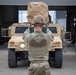 A U.S. Army soldier in OCP (Operational Camouflage Pattern) uniform is giving hand signals with both thumbs up while guiding a tan HMMWV (Humvee) military vehicle. The scene appears to take place at a military installation or motor pool, with a large hangar or garage structure visible in the background. At least two Humvees are visible, with one bearing a covered load marked 'FRONT 1' on top.