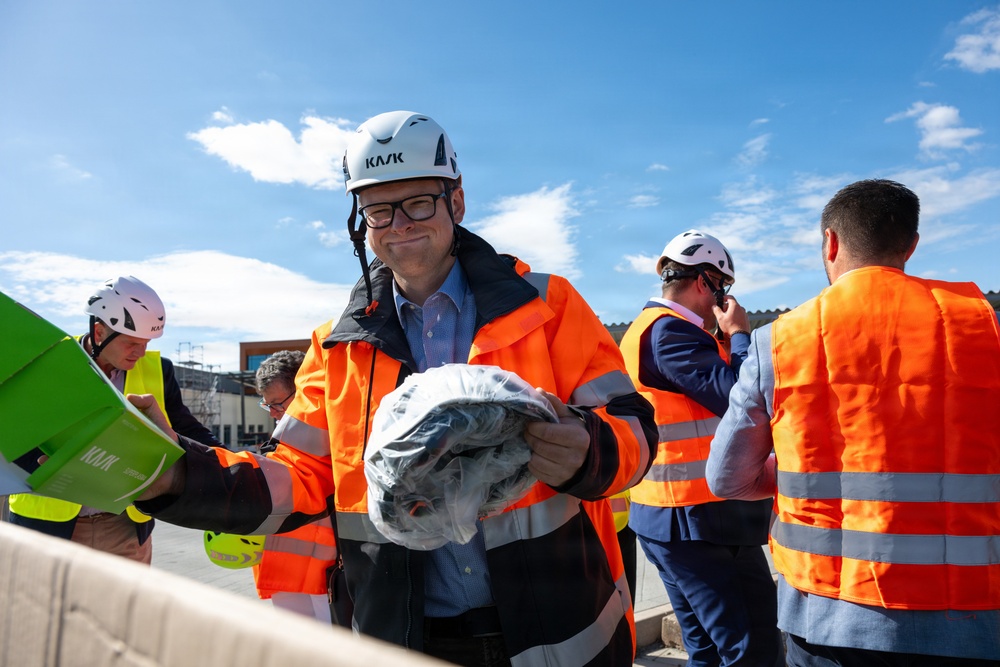 A group of individuals wearing orange high-visibility safety vests and white KASK brand hard hats are gathered at what appears to be a construction or industrial site. The central figure, a man wearing glasses, is smiling and holding a plastic-wrapped item, possibly safety equipment being distributed. The setting is outdoors under a partly cloudy blue sky.