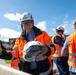 A group of individuals wearing orange high-visibility safety vests and white KASK brand hard hats are gathered at what appears to be a construction or industrial site. The central figure, a man wearing glasses, is smiling and holding a plastic-wrapped item, possibly safety equipment being distributed. The setting is outdoors under a partly cloudy blue sky.