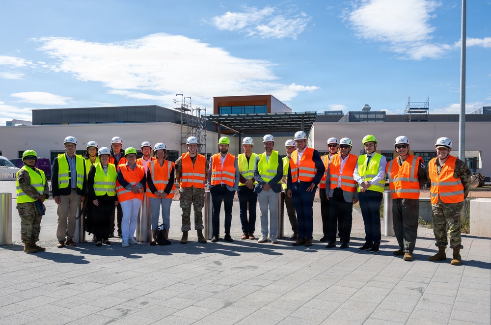 A group of approximately 18-20 individuals are posed for a group photograph at what appears to be an active construction or development site. The group includes a mix of civilians, military personnel, and site visitors, all wearing personal protective equipment including hard hats and high-visibility vests. The setting appears to be outside a modern building under construction or renovation.