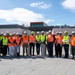 A group of approximately 18-20 individuals are posed for a group photograph at what appears to be an active construction or development site. The group includes a mix of civilians, military personnel, and site visitors, all wearing personal protective equipment including hard hats and high-visibility vests. The setting appears to be outside a modern building under construction or renovation.