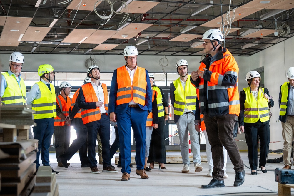 A group of approximately 10-12 people are conducting a site visit or inspection tour inside a building under construction. Participants are wearing safety equipment including white hard hats branded 'KAJI' and high-visibility vests in orange and yellow. The interior space shows an unfinished ceiling with exposed wiring, conduits, and structural elements.