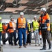 A group of approximately 10-12 people are conducting a site visit or inspection tour inside a building under construction. Participants are wearing safety equipment including white hard hats branded 'KAJI' and high-visibility vests in orange and yellow. The interior space shows an unfinished ceiling with exposed wiring, conduits, and structural elements.