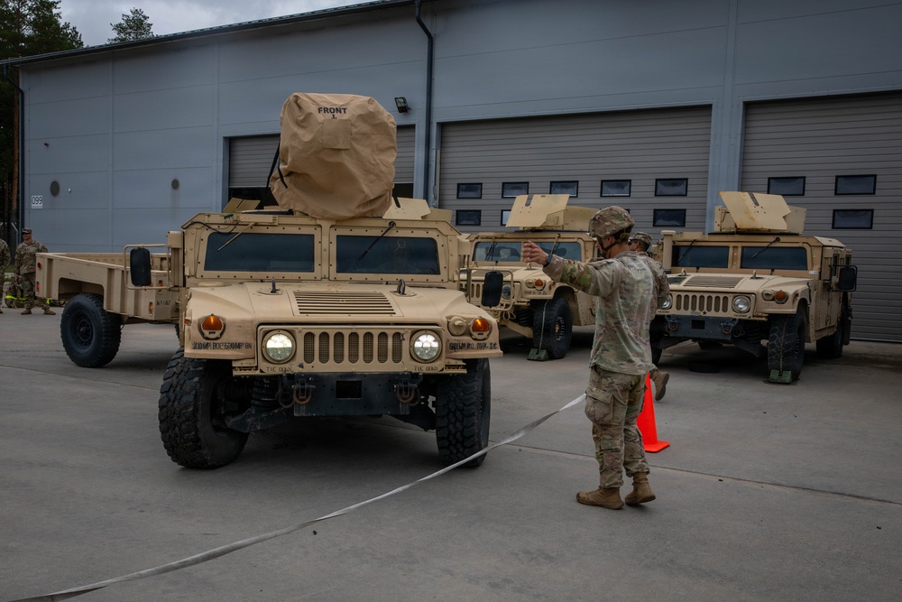 A military vehicle staging area showing multiple tan/desert-colored HMMWVs (Humvees) parked in front of a large building with garage doors. A soldier in camouflage uniform is directing or signaling toward the vehicles, with an orange traffic cone visible nearby. The turret-mounted equipment on the vehicles is covered with tan protective covers labeled 'FRONT'.