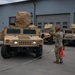 A military vehicle staging area showing multiple tan/desert-colored HMMWVs (Humvees) parked in front of a large building with garage doors. A soldier in camouflage uniform is directing or signaling toward the vehicles, with an orange traffic cone visible nearby. The turret-mounted equipment on the vehicles is covered with tan protective covers labeled 'FRONT'.