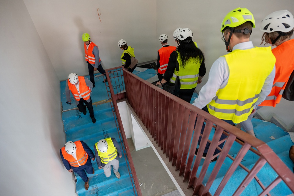 A group of approximately 9-10 people wearing safety helmets and high-visibility vests are conducting a site inspection or tour of a building under construction. They are navigating a stairwell with blue protective floor covering and a red metal railing. The photo is taken from an elevated angle looking down into the stairwell.