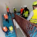 A group of approximately 9-10 people wearing safety helmets and high-visibility vests are conducting a site inspection or tour of a building under construction. They are navigating a stairwell with blue protective floor covering and a red metal railing. The photo is taken from an elevated angle looking down into the stairwell.
