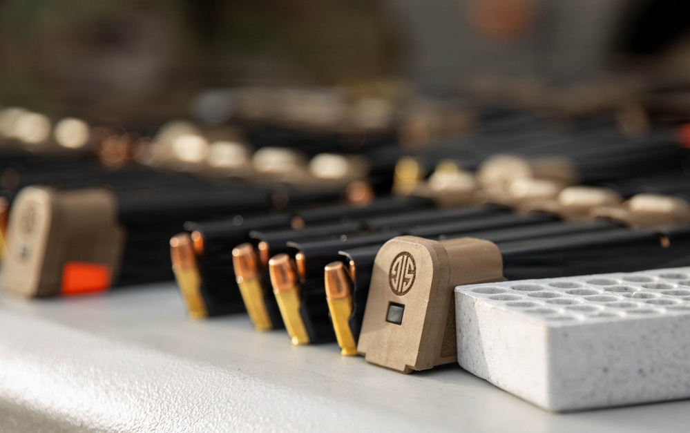 The image shows a close-up of firearm magazines, loose ammunition rounds, and a SIG Sauer branded magazine base plate arranged on a flat surface. Multiple loaded and unloaded magazines are visible in the background, along with what appears to be an ammunition loading tray on the right. The scene appears to be a shooting range or firearms preparation area.