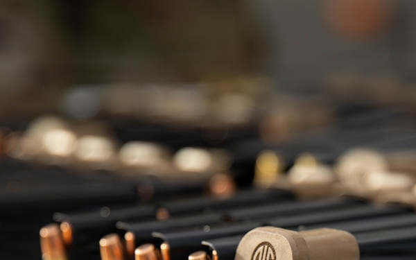 The image shows a close-up of firearm magazines, loose ammunition rounds, and a SIG Sauer branded magazine base plate arranged on a flat surface. Multiple loaded and unloaded magazines are visible in the background, along with what appears to be an ammunition loading tray on the right. The scene appears to be a shooting range or firearms preparation area.
