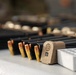 The image shows a close-up of firearm magazines, loose ammunition rounds, and a SIG Sauer branded magazine base plate arranged on a flat surface. Multiple loaded and unloaded magazines are visible in the background, along with what appears to be an ammunition loading tray on the right. The scene appears to be a shooting range or firearms preparation area.