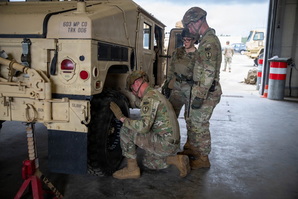 Three U.S. Army Military Police soldiers are performing maintenance on a HMMWV (Humvee) vehicle, specifically working on the front tire. The vehicle is marked '603 MP CO TRK 006', identifying it as belonging to the 603rd Military Police Company. The scene takes place in what appears to be a motor pool or maintenance bay.