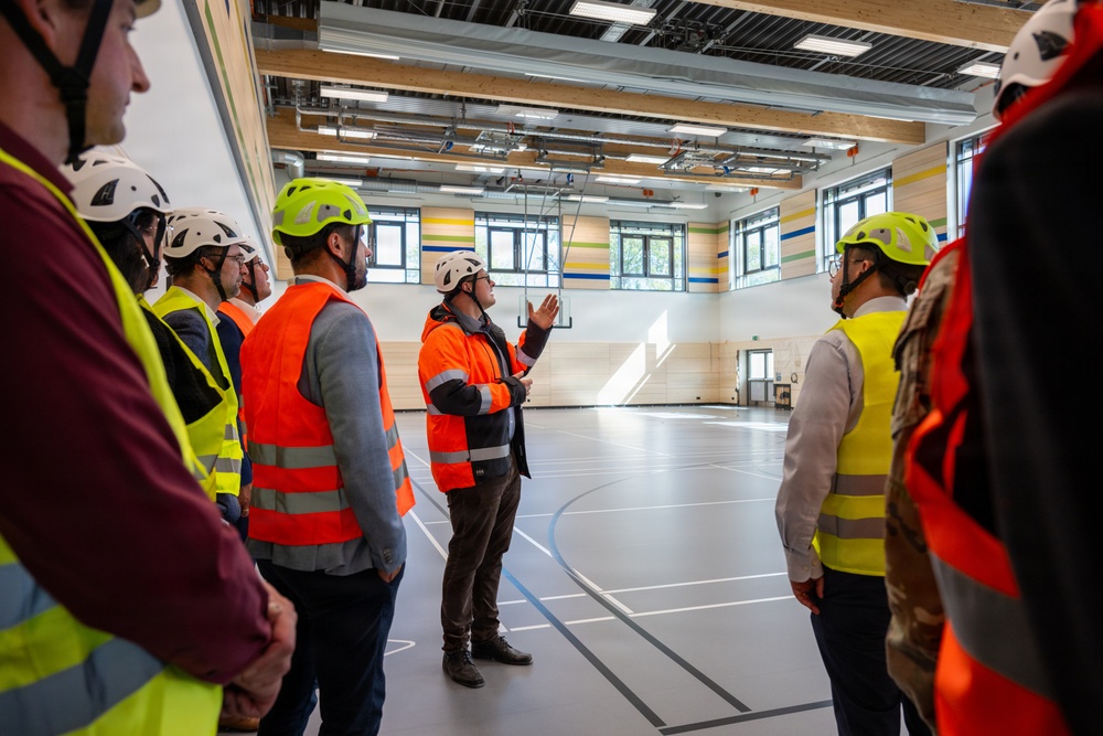 A group of individuals wearing high-visibility safety vests and hard hats are conducting a site inspection or guided tour inside a newly constructed or renovated gymnasium/sports hall. One person in an orange vest and white helmet appears to be leading the group, gesturing upward toward the ceiling infrastructure. The facility features a clean sports floor with court markings, exposed ceiling systems, and colorful wall striping.