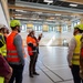A group of individuals wearing high-visibility safety vests and hard hats are conducting a site inspection or guided tour inside a newly constructed or renovated gymnasium/sports hall. One person in an orange vest and white helmet appears to be leading the group, gesturing upward toward the ceiling infrastructure. The facility features a clean sports floor with court markings, exposed ceiling systems, and colorful wall striping.