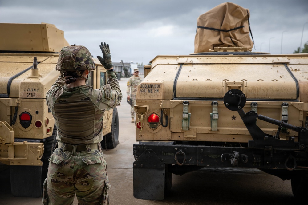 A U.S. Army soldier in full combat gear is directing military vehicle movement, raising one hand as a ground guide signal between two tan HMMWV (Humvee) vehicles. The vehicles are marked with unit identifiers '300 MP BDE / 530 MP BN', indicating Military Police Brigade and Battalion affiliation. The scene appears to take place on a paved staging area under overcast skies.