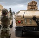 A U.S. Army soldier in full combat gear is directing military vehicle movement, raising one hand as a ground guide signal between two tan HMMWV (Humvee) vehicles. The vehicles are marked with unit identifiers '300 MP BDE / 530 MP BN', indicating Military Police Brigade and Battalion affiliation. The scene appears to take place on a paved staging area under overcast skies.