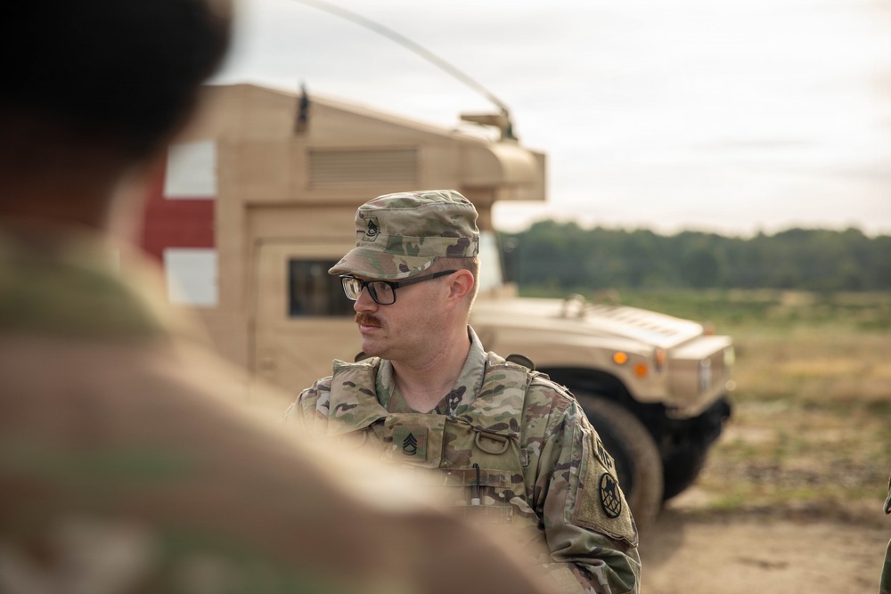 A U.S. Army soldier wearing Operational Camouflage Pattern (OCP) uniform and glasses is standing near a tan military HMMWV (Humvee) marked with a red cross, indicating a medical vehicle. The soldier appears to be a Staff Sergeant based on the rank insignia visible on the patrol cap. The setting appears to be an outdoor training or operational environment with open terrain and tree line in the background.