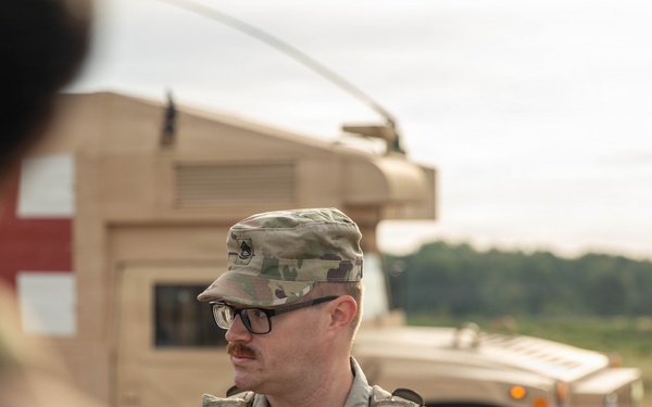 A U.S. Army soldier wearing Operational Camouflage Pattern (OCP) uniform and glasses is standing near a tan military HMMWV (Humvee) marked with a red cross, indicating a medical vehicle. The soldier appears to be a Staff Sergeant based on the rank insignia visible on the patrol cap. The setting appears to be an outdoor training or operational environment with open terrain and tree line in the background.