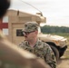 A U.S. Army soldier wearing Operational Camouflage Pattern (OCP) uniform and glasses is standing near a tan military HMMWV (Humvee) marked with a red cross, indicating a medical vehicle. The soldier appears to be a Staff Sergeant based on the rank insignia visible on the patrol cap. The setting appears to be an outdoor training or operational environment with open terrain and tree line in the background.