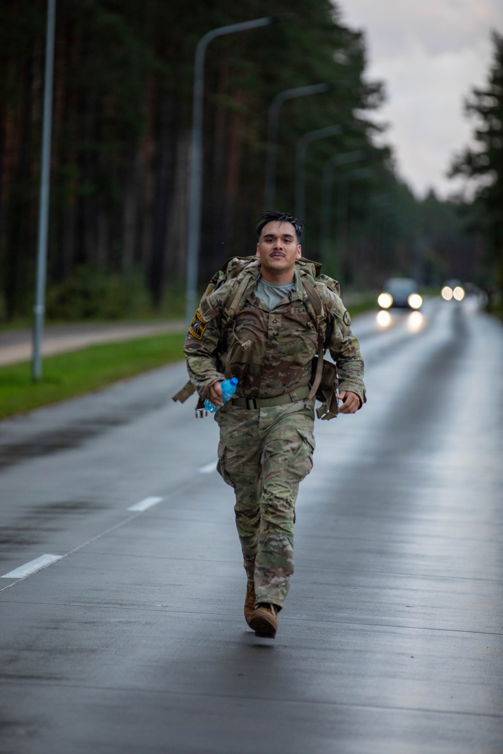 A U.S. Army soldier in full OCP (Operational Camouflage Pattern) uniform is marching or rucking along a wet, tree-lined road while carrying a loaded rucksack and a water bottle. The road appears to be on a military installation or training area, with street lamps and vehicles visible in the background. The overcast, damp conditions suggest this is an endurance or physical fitness event such as a ruck march.