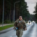 A U.S. Army soldier in full OCP (Operational Camouflage Pattern) uniform is marching or rucking along a wet, tree-lined road while carrying a loaded rucksack and a water bottle. The road appears to be on a military installation or training area, with street lamps and vehicles visible in the background. The overcast, damp conditions suggest this is an endurance or physical fitness event such as a ruck march.