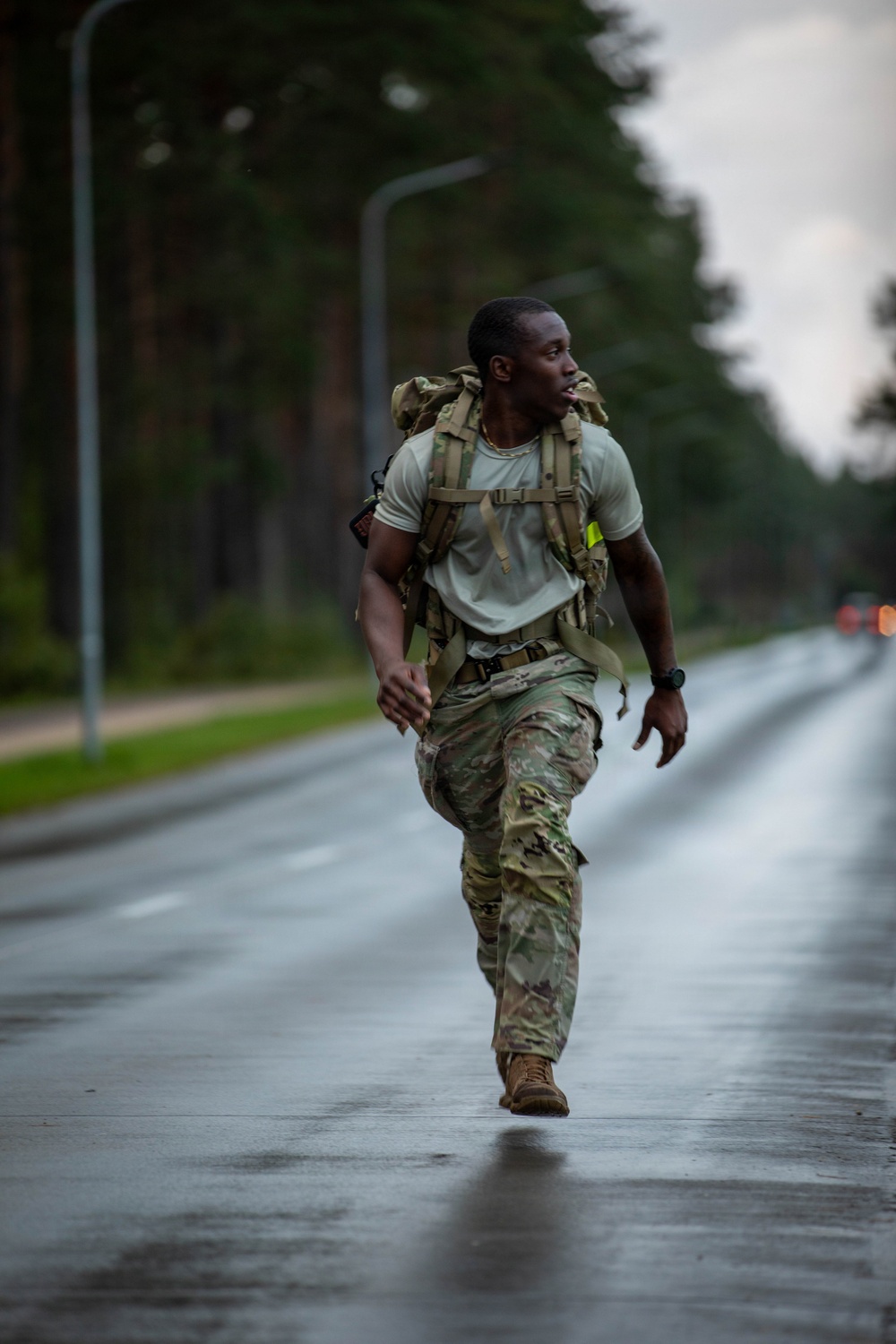 A soldier in military camouflage uniform is running along a wet road lined with tall trees and street lamps, carrying a large tactical rucksack on his back. The image appears to capture a military ruck march or physical training exercise under overcast, rainy conditions. The soldier appears physically exerted, with mouth open and body leaning forward in motion.