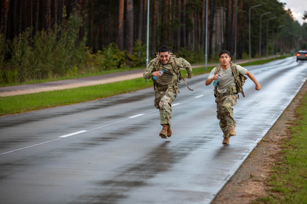Two military personnel in combat uniforms and rucksacks are running at full sprint along a wet road flanked by tall pine trees. Both soldiers are carrying water bottles and wearing tactical gear, suggesting they are participating in a ruck march or physical fitness event. The lead soldier is visibly smiling, indicating a competitive or celebratory finish.