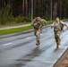 Two military personnel in combat uniforms and rucksacks are running at full sprint along a wet road flanked by tall pine trees. Both soldiers are carrying water bottles and wearing tactical gear, suggesting they are participating in a ruck march or physical fitness event. The lead soldier is visibly smiling, indicating a competitive or celebratory finish.