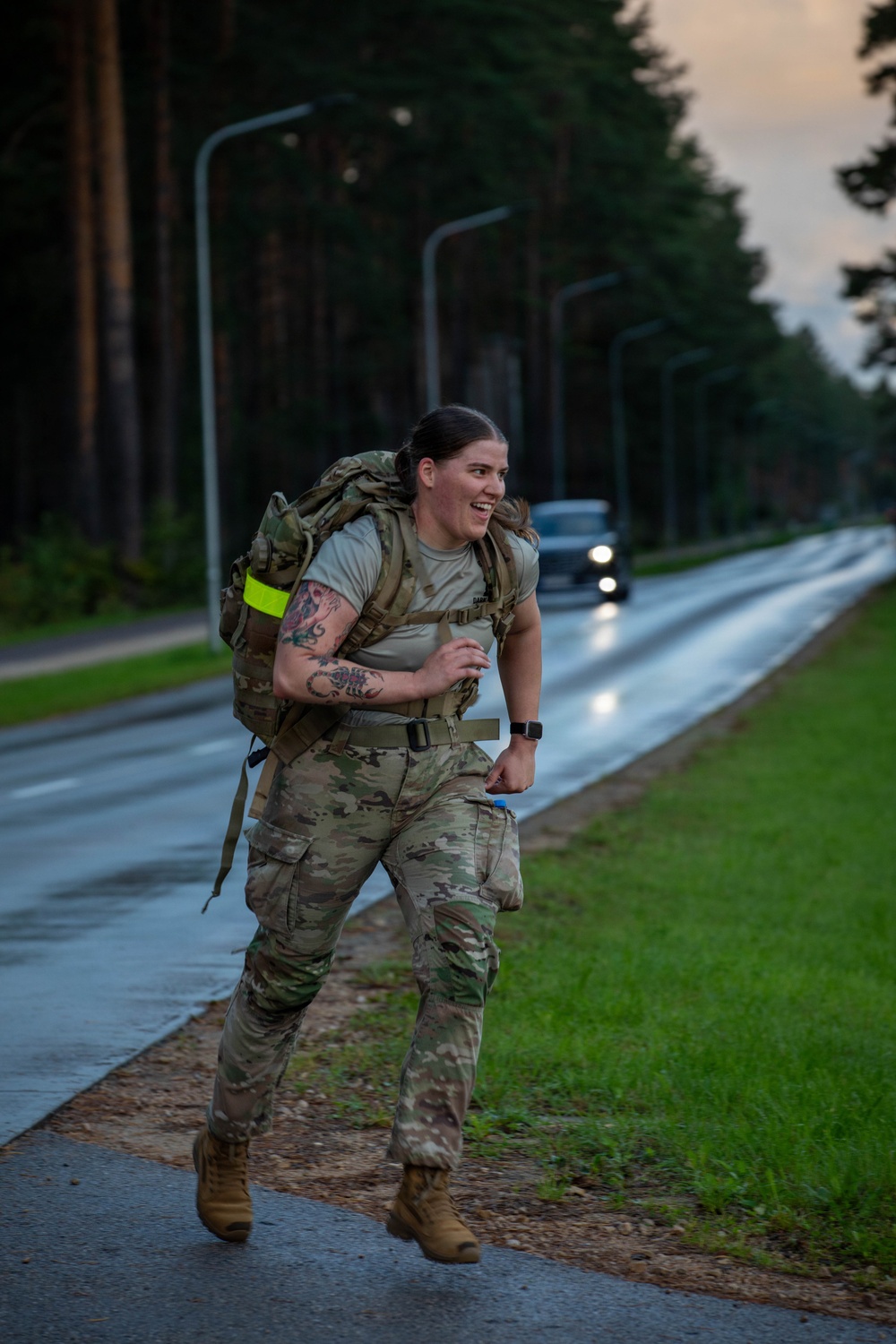 A female soldier in full military camouflage uniform (OCP/MultiCam pattern) is conducting a ruck march along a paved road at dusk or dawn. She is carrying a large tactical rucksack with a high-visibility yellow safety band attached, and appears to be moving at a brisk pace with a smile on her face. The setting appears to be a military installation road lined with tall trees and street lamps.