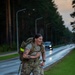 A female soldier in full military camouflage uniform (OCP/MultiCam pattern) is conducting a ruck march along a paved road at dusk or dawn. She is carrying a large tactical rucksack with a high-visibility yellow safety band attached, and appears to be moving at a brisk pace with a smile on her face. The setting appears to be a military installation road lined with tall trees and street lamps.